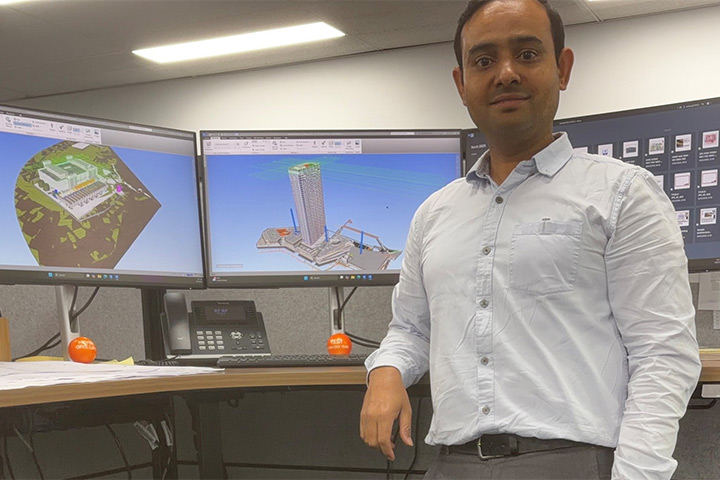 Vishu Kaushik stands facing the camera, wearing a button-up shirt and leaning his elbow on a stand-up office desk. There are three monitors on the desk showing technical images of a large construction project.