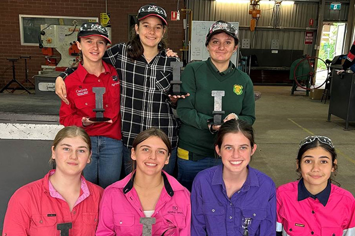 Seven girls from Carinya Christian School wearing work shirts, show off their handywork inside a large workshop. 