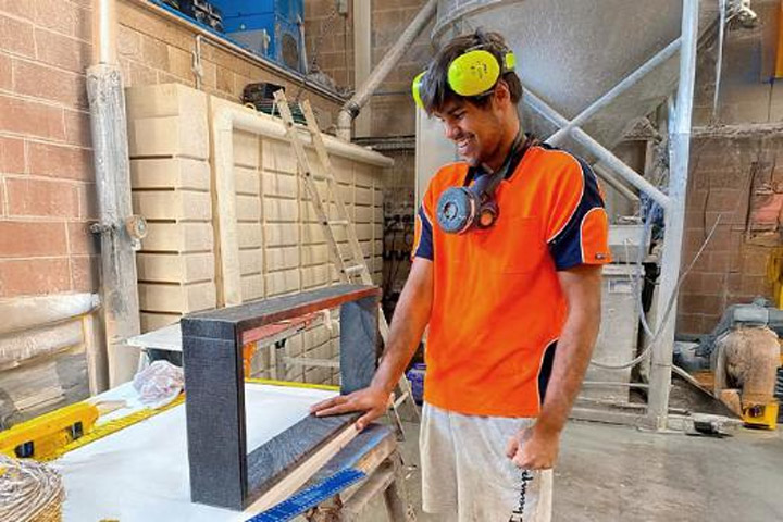 Shane McKay standing in a workshop wearing a bright orange t-shirt, earmuffs around his neck and a mask on his head. 