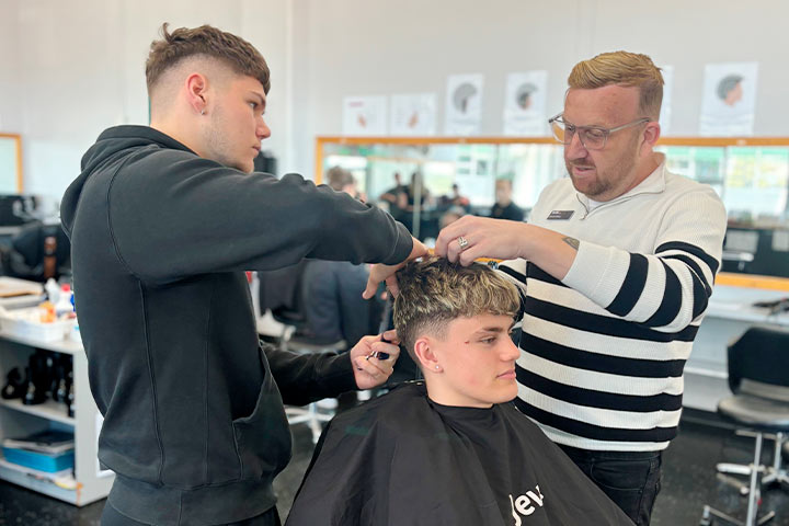 A TAFE NSW hairdressing teacher guides a student as he cuts a young man's hair in a salon classroom.