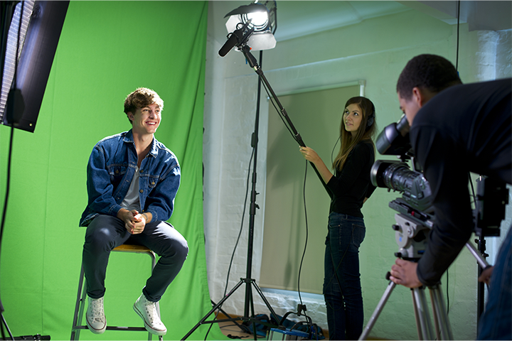 A male sits on a stool in front of a green screen, while a women holds a studio microphone and a man stands behind a camera.