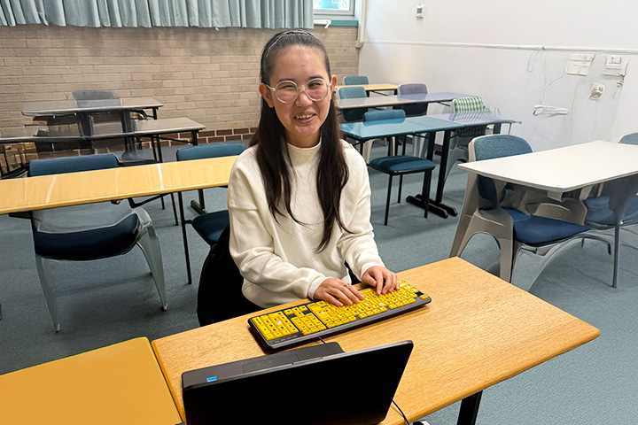 Samira Haidary is sitting at a desk in an empty classroom. She has long black hair and is wearing a white long sleeved top and glasses.