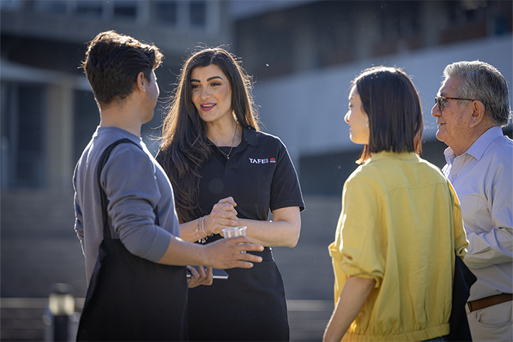 3 people stand outside, looking at a TAFE NSW staff member who is talking to them.