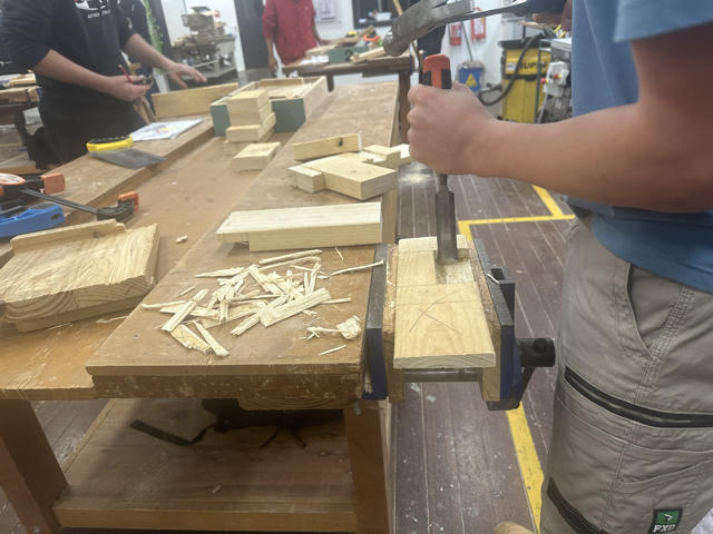 A group of students woodworking with hammers, chisels and other hand tools at a carpentry bench in a workshop.