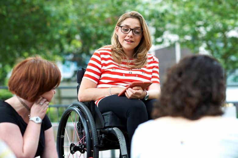 A woman in a wheelchair is holding a tablet in an outdoor setting. She has glasses on and wears a red and white striped top, while engaging in a conversation with two people seated in front of her.