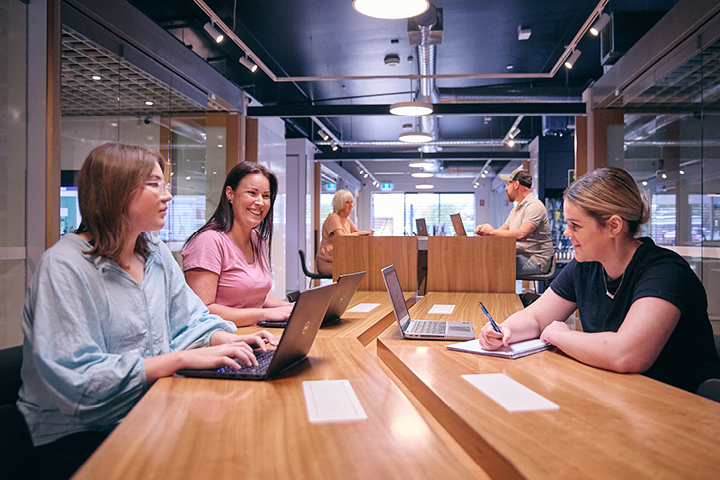 Three individuals smiling, working together on laptops in a well-lit learning centre.