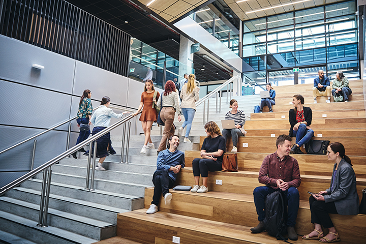 A modern TAFE NSW campus atrium that has a concrete set of stairs for people to walk up and down, and an adjacent wooden stair fixture that also serves as seating. There are people in pairs and singles sitting on steps talking and working.