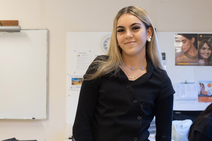Aliarna Dixon, a young woman with long blonde hair and light skin, smiles confidently in a classroom. She wears a black top, pearl earrings, and layered necklaces.