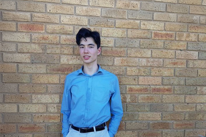 Brendan Bates, a graduate of TAFE Digital, smiles with his hands in his pockets. He is wearing a collared shirt and dress pants and stands outdoors in front of a beige and rust coloured brick wall.