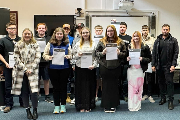 A group of eleven smiling teenage graduates of the Certificate II in Career Preparation at TAFE NSW Bathurst, standing in two rows, proudly holding their certificates, flanked by their TAFE NSW teachers.