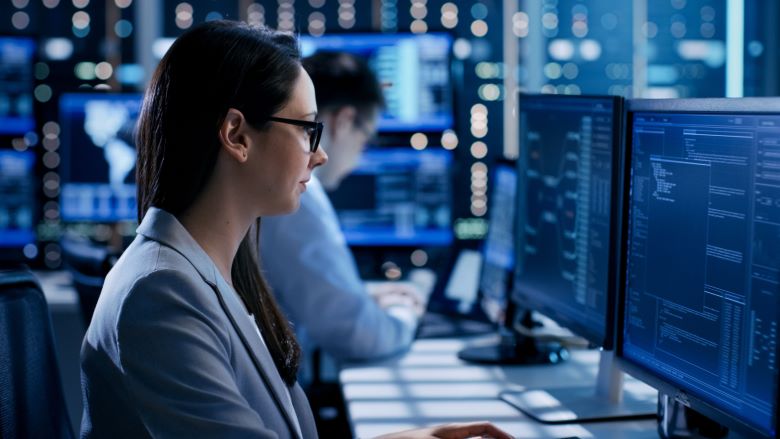 A woman looks at two large computer screens with code on them. She is in a computer lab surrounded by screens and technology, and wears glasses and a business suit.