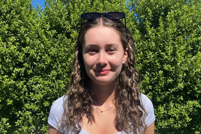 A photo of Josephine Bradbury smiling and standing outdoors in front of large green shrubbery.