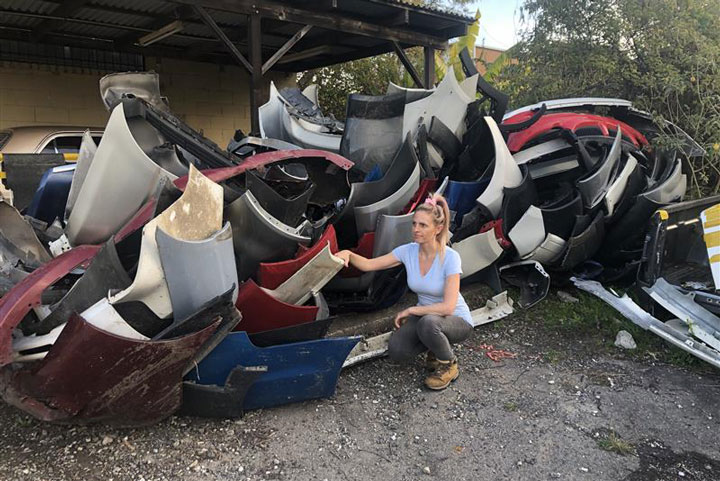 Rebecca Healy, a woman with blonde hair tied in a high ponytail, wearing a light blue T-shirt and dark pants, crouching beside a large pile of discarded plastic car bumper bars collected for recycling, outside a shed.