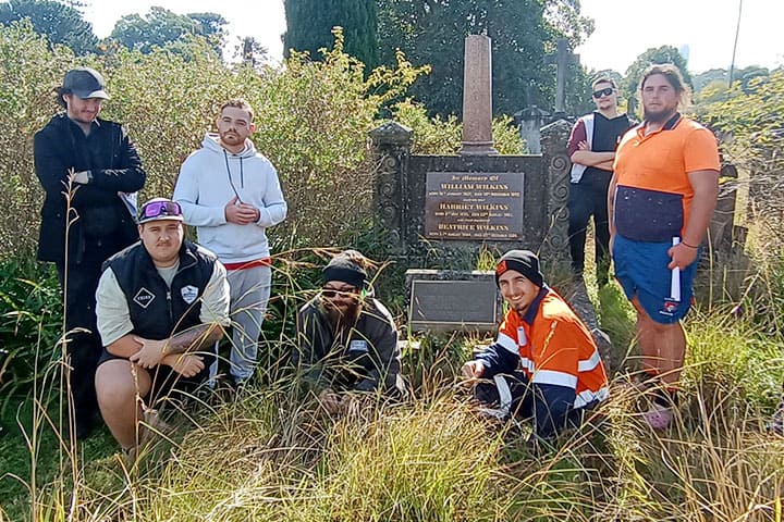Seven TAFE NSW stonemasonry apprentices at Rookwood Cemetery pictured next to an old headstone surrounded by tall grass.