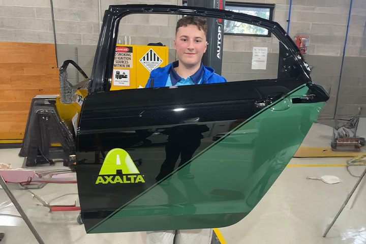 Samuel Shedden standing behind a freshly painted car door in a workshop. The door features a black and green design with the Axalta logo.