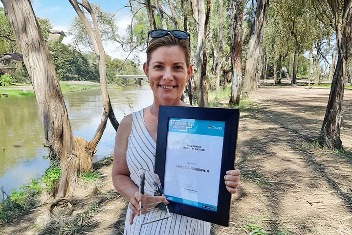 Tamileigh Chirgwin smiling and holding a framed award and trophy beside a riverbank lined with tall trees. The award reads: “TAFE NSW Excellence Awards – Environmental Sustainability Student of the Year – Tamileigh Chirgwin.”