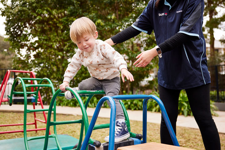 A young child with blonde hair is climbing on a colourful jungle gym outdoors. An adult, dressed in a navy TAFE NSW shirt with 'Childhood' written on it, is standing nearby, providing support.