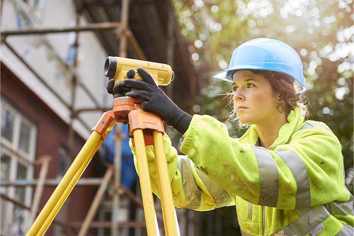 Female construction work stands at a construction site looking through a device on a tripod.