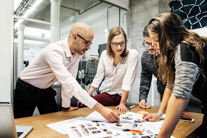 A diverse group of four professionals gather around a table in a modern office space. The table is covered with documents, fashion images and photos. They are physically engaging in the material while discussing ideas.