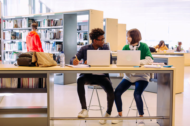Two casually dressed students sit at a study-bench in a modern library reviewing TAFE NSW policies. They both have open laptops in front of them. One student smiles to the other who appears to be sharing ideas with him. Other students are in the background studying and perusing the bookshelves.