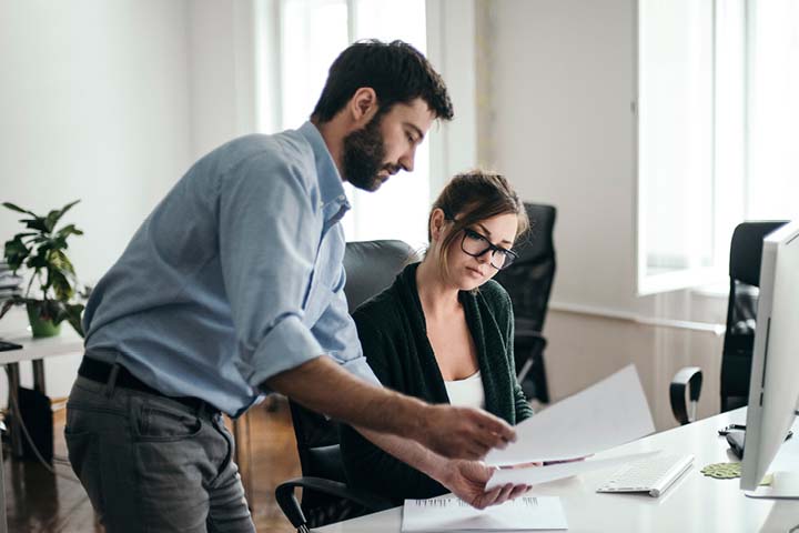 Two people in an office looking at documents, one seated at a desk. 