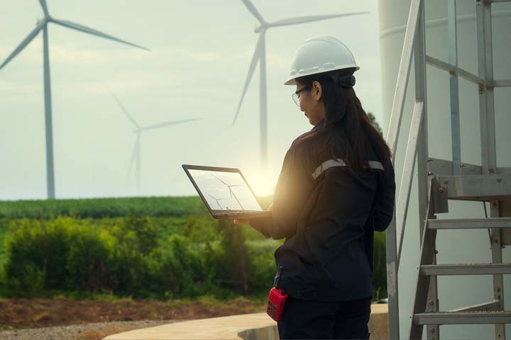 Wind energy engineer, female, working remotely on laptop with wind turbines in the distance.
