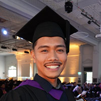 Person wearing a graduation cap and gown standing indoors with other people in the background.