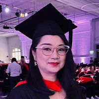Person wearing a black graduation cap and gown with a red hood, standing in an indoor venue with other people in graduation attire in the background.