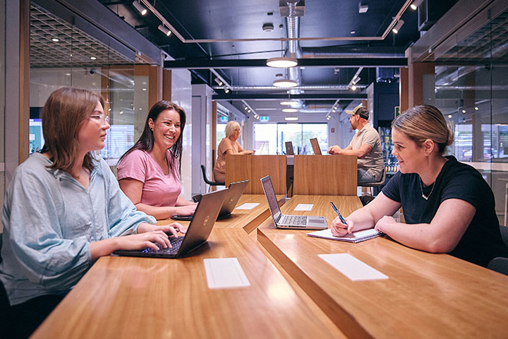 Three people participate in a boardroom meeting at TAFE NSW, seated at modern wooden tables with laptops and notebooks, engaging in collaborative discussions in a well-lit room with glass partitions.