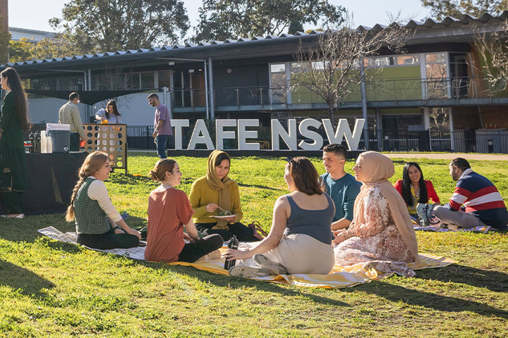 People sit on blankets on a grassy area at a TAFE NSW campus, enjoying an outdoor BBQ with games and social activities, surrounded by trees and buildings, promoting community engagement.
