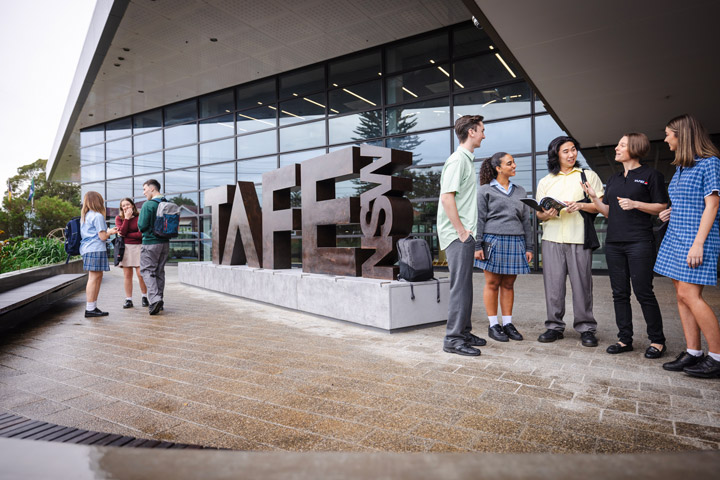 Three students with backpacks engage with a TAFE NSW staff member at an on-campus information booth, featuring a display of vocational training in technical fields like solar energy and industrial equipment.