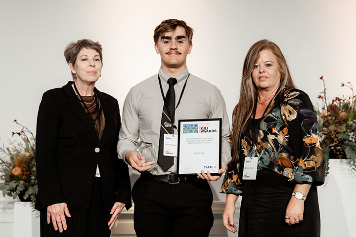 Three people standing together, with the person in the middle holding a framed certificate and a glass award.  The individuals on either side are in formal attire with name tags, and floral arrangements are visible in the background.