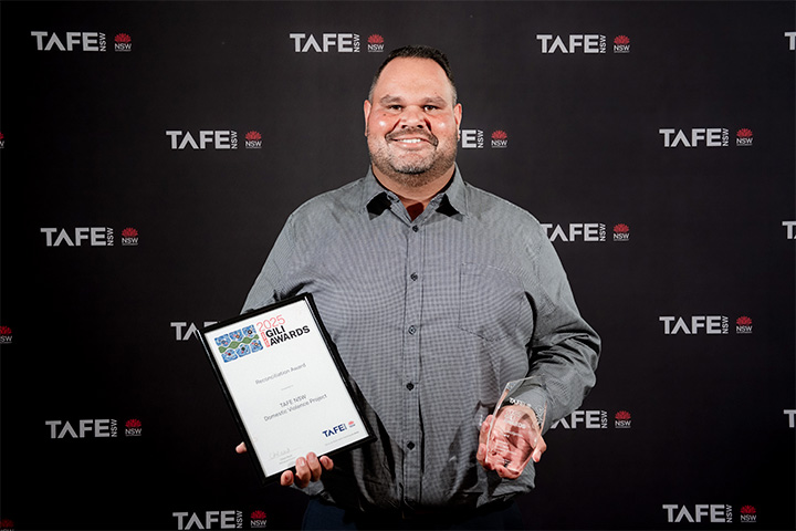 A person holding a framed certificate and a glass award in front of a TAFE NSW backdrop. The certificate reads: “2025 Gili Awards – Domestic Violence Project.”