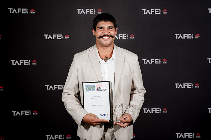 A person in a light-colored suit holding a certificate and an award in front of a TAFE NSW backdrop. The certificate reads: “2025 Gili Awards – Lachlan Peachey.”