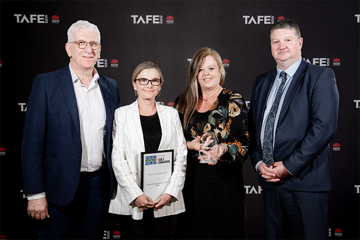 Four individuals standing in front of a TAFE NSW backdrop. One holds a framed certificate reading “2025 Gili Awards – Precise Air Aboriginal Pathways,” and another holds a glass award.