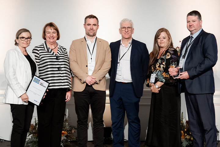 Six people standing in a row in formal attire. One person on the left holds a certificate or award, and another on the right holds a glass trophy. The background features a plain white wall with floral decorations at the bottom.