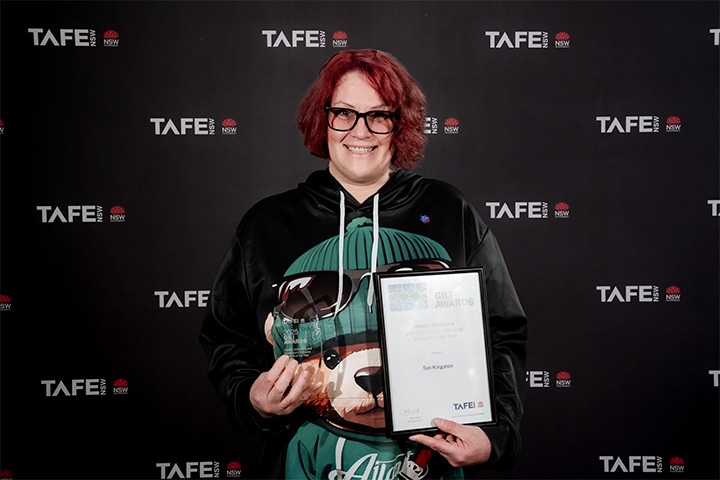 A person holding an award and a certificate in front of a TAFE NSW backdrop. The certificate reads: “2025 Gili Awards – Tori Kingston.”