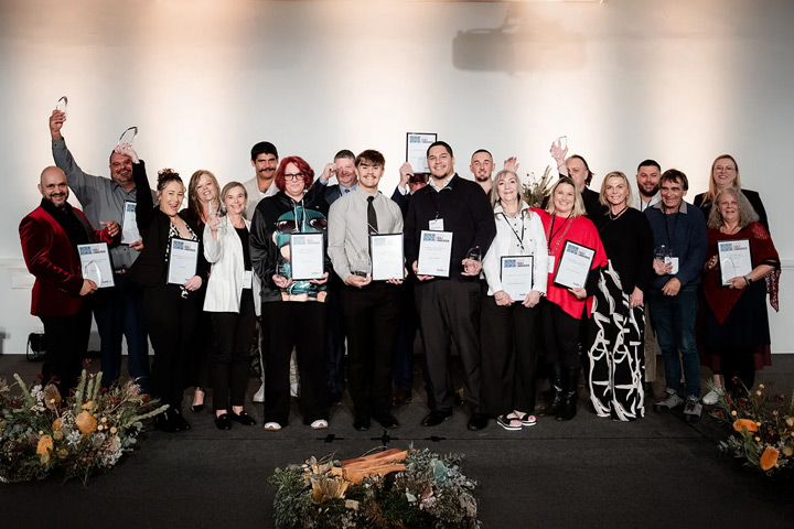 Group of individuals standing on a stage holding awards and certificates, with floral arrangements in front and a softly lit background.