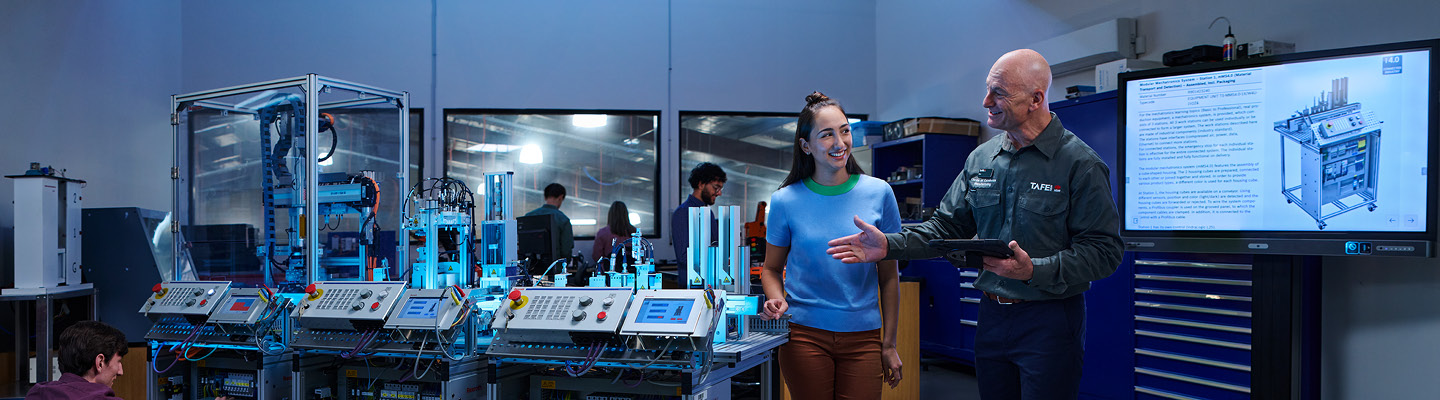 TAFE NSW instructor holding a tablet and explaining a concept to a student in a workshop setting with a technical display in the background