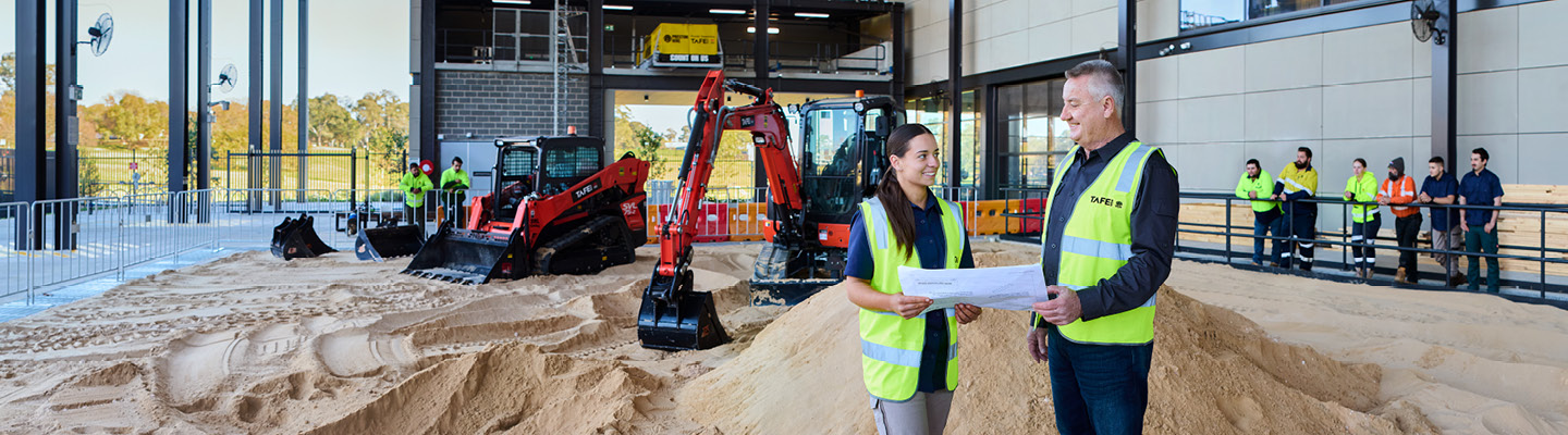 Student and teacher in a civil construction class at a simulated work site, excavator in background.