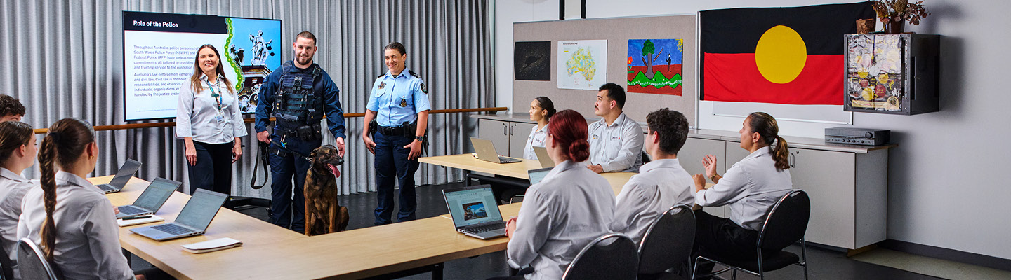 Classroom scene with students using laptops, and three presenters at the front including a police officer, a tactical officer with a dog, and a speaker in a white shirt, with a screen displaying 'Role of the Police'