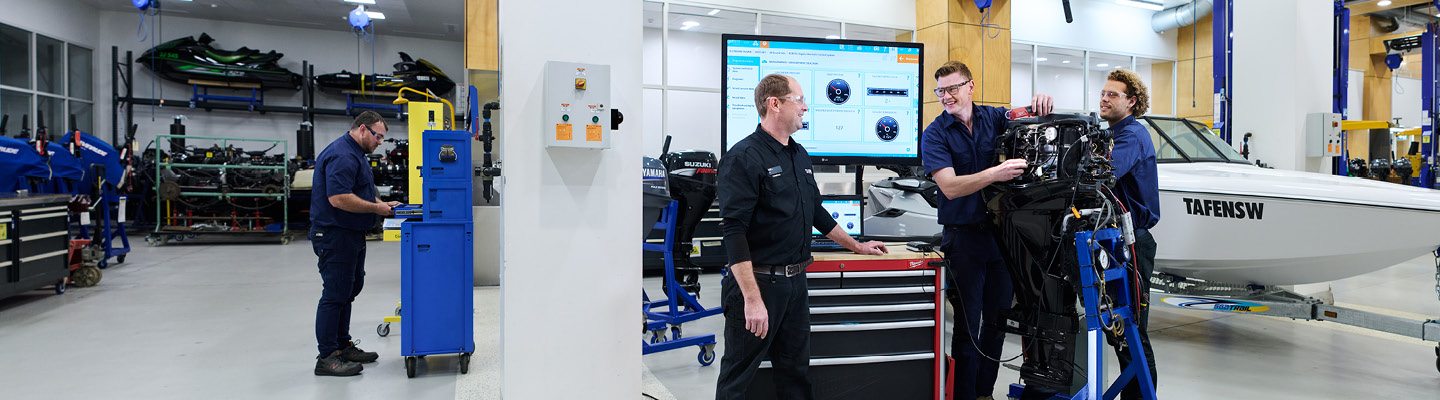 A male marine mechanic in a classroom workshop setting, guiding two students as they work together to troubleshoot and repair an outboard. A screen with instructional materials is visible in the background