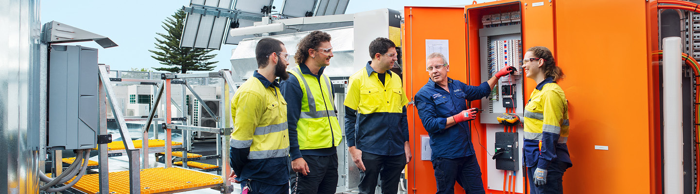A workshop scene with an electrotechnology teacher guiding four students as they collaborate on a hands-on electrical project.