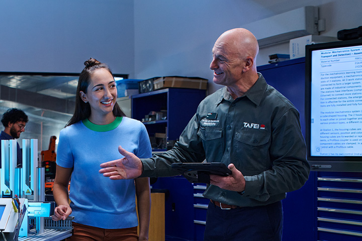 TAFE NSW instructor holding a tablet and explaining a concept to a student in a workshop setting with a technical display in the background
