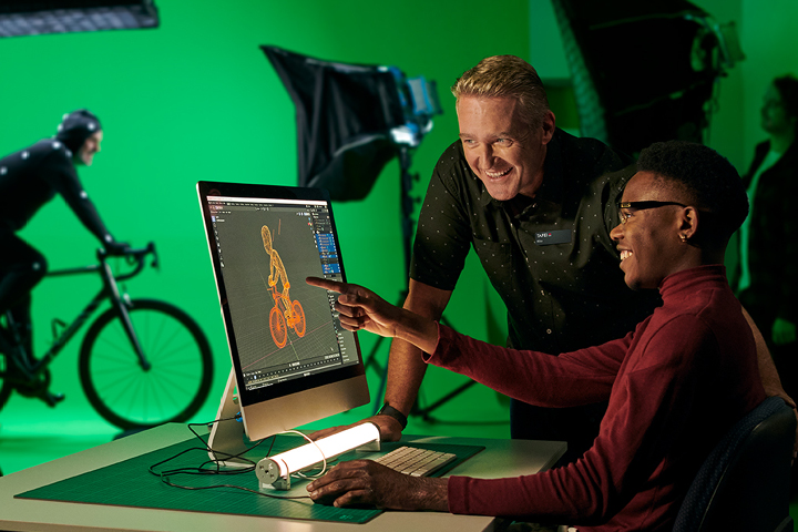 A young male designer works on a graphic design project on his computer in a brightly lit design studio. His teacher stands beside him offering guidance. In the background, a photographer shoots a scene involving a man riding a bicycle in front of a green screen.