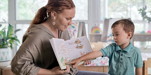 A woman reading with a young girl who is pointing at a book, and they are in a classroom setting.