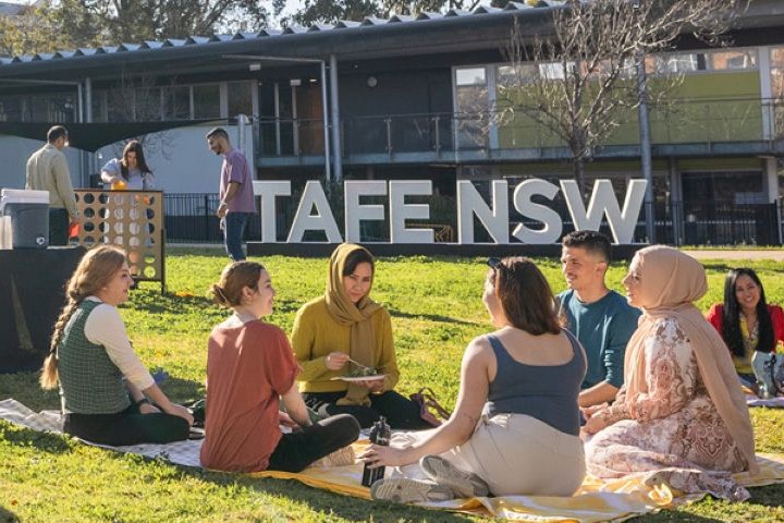 People sit on blankets on a grassy area at a TAFE NSW campus, enjoying an outdoor BBQ with games and social activities, surrounded by trees and buildings, promoting community engagement.