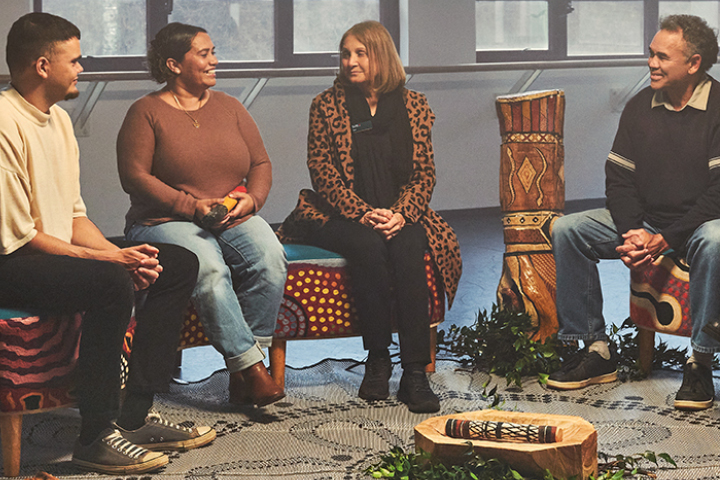 An Indigenous mob sitting in a yarning semi circle, engaging in conversation. There is a round rug that they are sitting on around, leaves, plants and other culturally significant pieces. 
