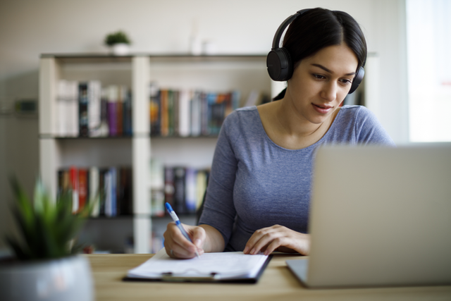 A woman sitting at a desk and writing notes, looking at her laptop and wearing headphones.