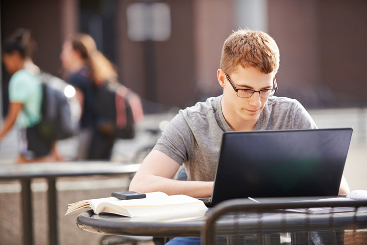 A young man wearing glasses sits in an outdoor area while working on his laptop.